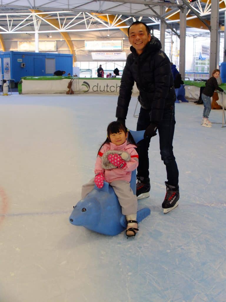 Foto van een vader die zijn dochter op een schaatshulp (dolfijntje) voortduwd op de ijsbaan 